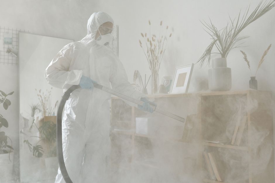 A professional cleaner from Fulham Carpet Cleaners is performing surface cleaning in a bright, modern living room. The cleaner is wearing full protective gear, including a white coverall suit, safety