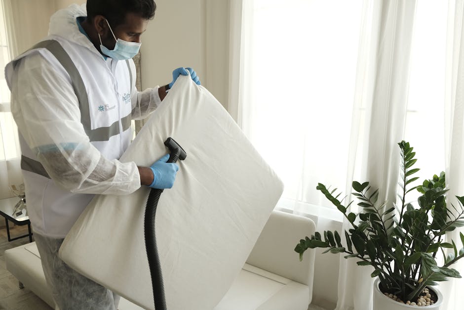 A professional cleaner from Fulham Carpet Cleaners is performing surface cleaning in a bright, modern living room. The cleaner is wearing full protective gear, including a white coverall suit, safety