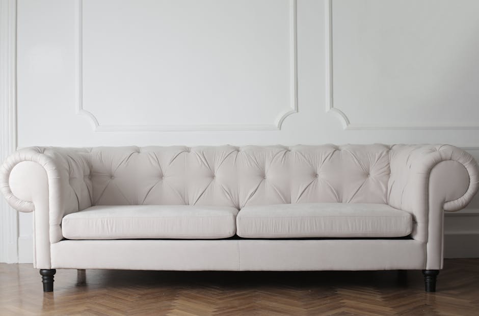 A close-up of a person's hand holding a vacuum cleaner nozzle, actively cleaning the olive-green upholstered armchair in a well-lit living room. Behind the armchair, a dark gray fabric sofa is visible